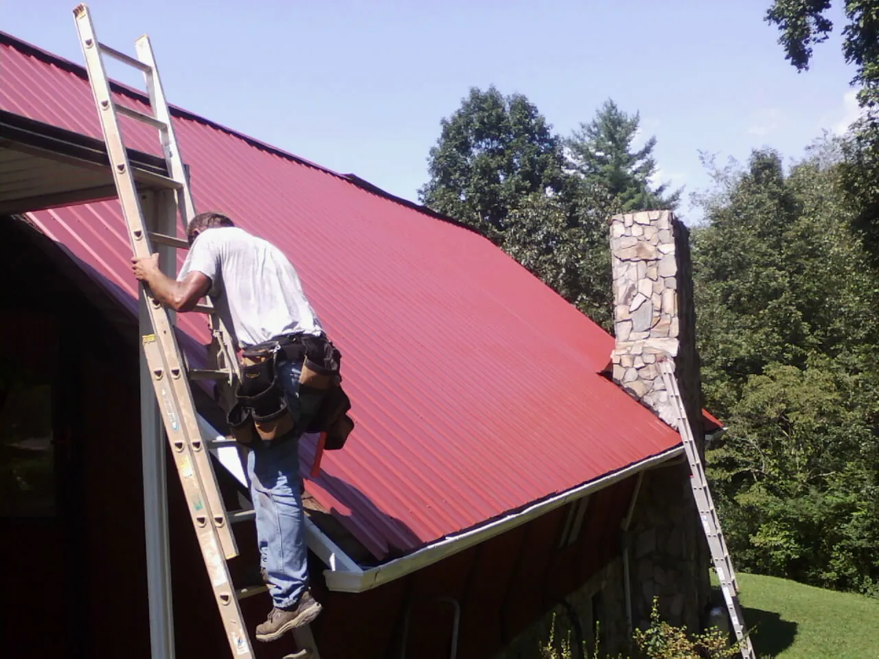 Roofer on a ladder beside a red metal roof and stone chimney — Soffit & Fascia Repair by Roofing Brothers in Parma